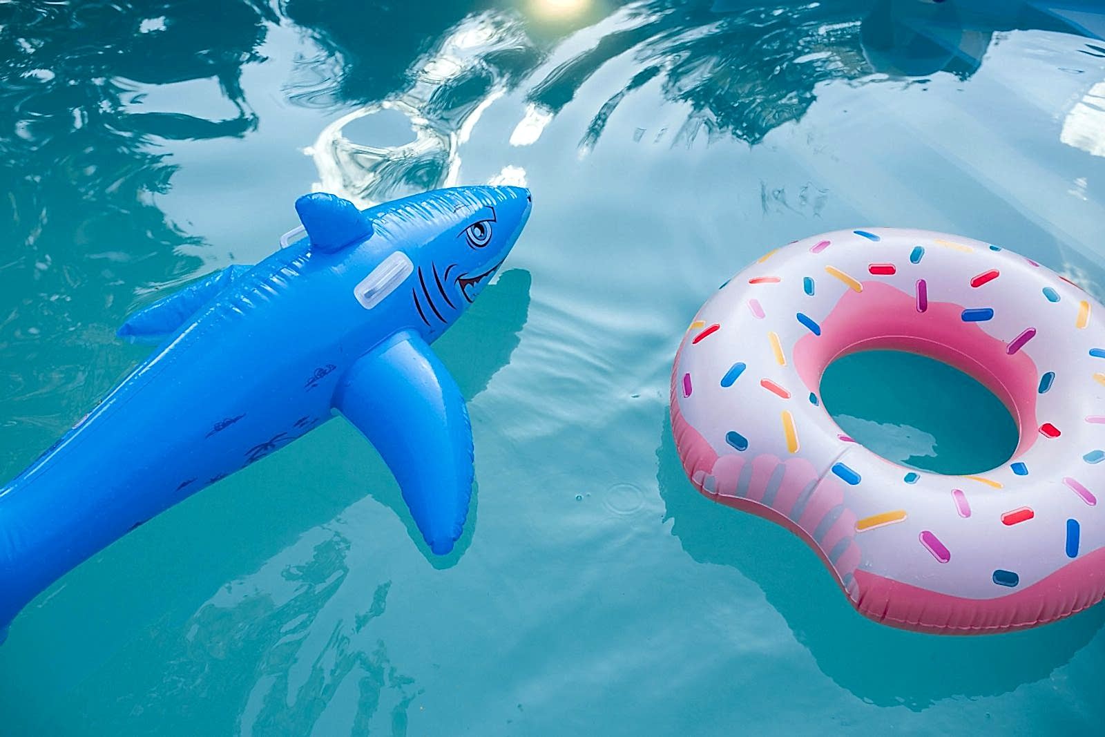 Brightly colored inflatable shark and donut floats in a sunlit swimming pool.